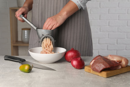 Woman making chicken mince with metal meat grinder at gray table indoors, closeup