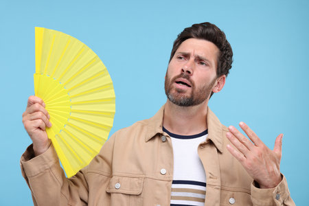 Unhappy man with hand fan suffering from heat on light blue backgroundの写真素材
