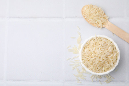 Bowl and spoon with raw rice on white tiled table, flat lay. Space for textの写真素材