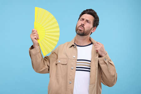 Unhappy man with hand fan suffering from heat on light blue backgroundの写真素材