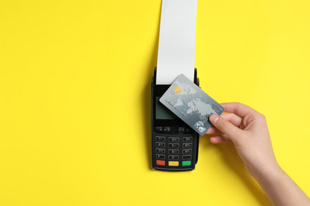 Woman using payment terminal with credit card and thermal paper for receipt on yellow background, top view. Space for textの写真素材