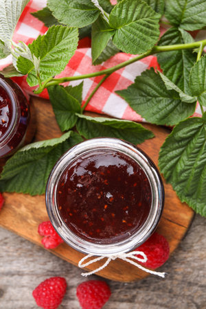 Delicious raspberry jam, fresh berries and green leaves on wooden table, flat lay