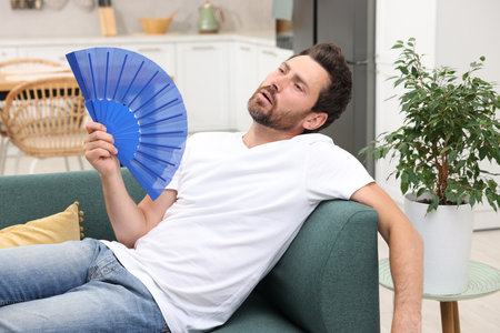 Bearded man waving blue hand fan to cool himself on sofa at homeの写真素材