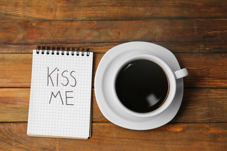 Notebook with phrase Kiss Me and cup of coffee on wooden table, flat layの写真素材