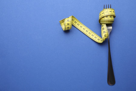 Fork with measuring tape on blue background, top view and space for text. Diet concept