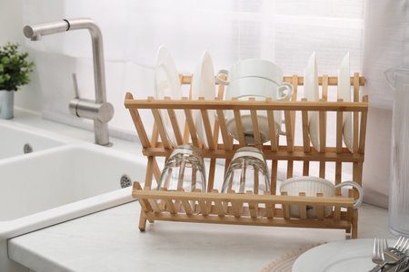 Drying rack with clean dishes on light marble countertop near sink in kitchen