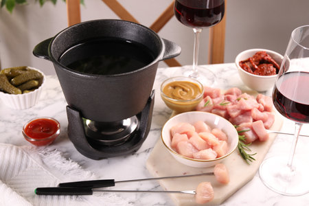 Fondue pot with oil, forks, raw meat pieces, glasses of red wine and other products on white marble tableの写真素材