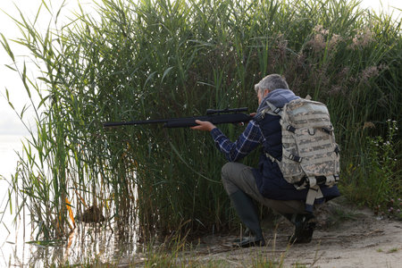 Man aiming with hunting rifle near lake outdoors. Space for textの写真素材