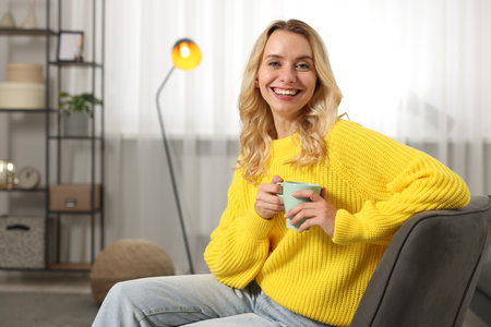 Happy woman in stylish warm sweater holding cup of drink at home. Space for textの写真素材