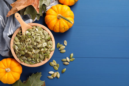 Bowl with seeds, fresh pumpkins and dry leaves on blue wooden table, flat lay. Space for text