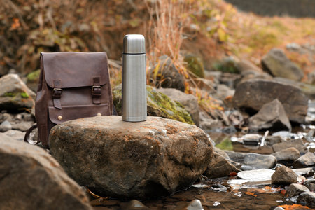 Metallic flask and leather backpack on stone near river, space for textの写真素材