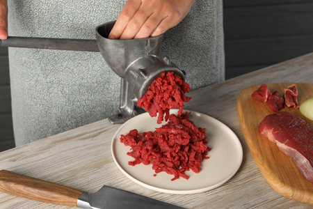 Woman making beef mince with manual meat grinder at light wooden table, closeup