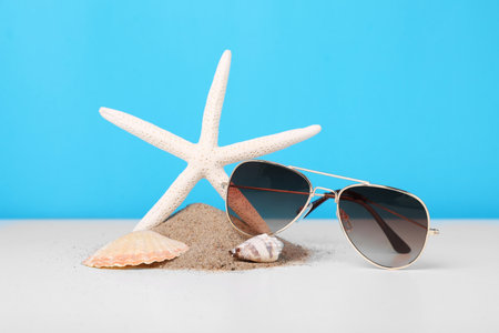 Stylish sunglasses, seashells, starfish and sand on white table against light blue backgroundの写真素材