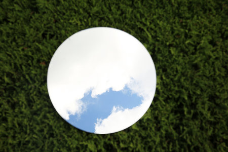 Round mirror on green grass reflecting cloudy sky, top viewの写真素材