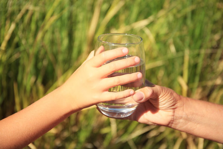 Child giving glass of water to elderly woman outdoors on sunny day, closeup