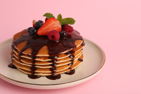 Stack of tasty pancakes with fresh berries, chocolate sauce and mint on pink background, closeup....