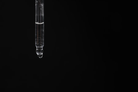 Dripping cosmetic serum from pipette against black background, space for textの写真素材