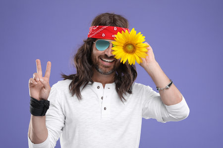 Stylish hippie man covering eye with sunflower and showing V-sign on violet backgroundの写真素材