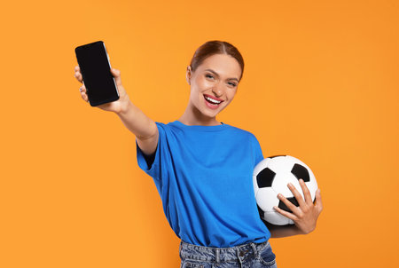 Happy fan with football ball showing smartphone on orange backgroundの写真素材