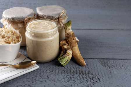 Spicy horseradish sauce in jars, roots and spoon on gray wooden table, space for textの写真素材