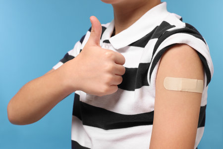 Boy with sticking plaster on arm after vaccination showing thumbs up against light blue background, closeupの写真素材