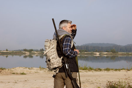Man with hunting rifle and backpack near lake outdoors