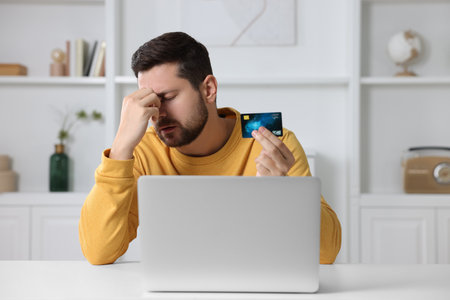 Upset man with credit card near laptop at home. Be careful - fraudの写真素材