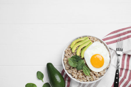 Tasty boiled oatmeal with fried egg, avocado and basil served on white wooden table, flat lay. Space for textの写真素材
