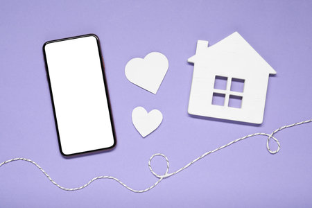 Long-distance relationship concept. Smartphone, white house, paper hearts and decorative cord on violet background, flat layの写真素材