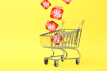 Discount offer. Gift boxes with percent signs falling into shopping trolley on yellow background, space for textの写真素材