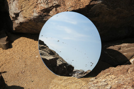Round mirror reflecting light blue sky on sand near stones outdoorsの写真素材