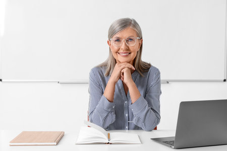 Happy professor sitting near laptop at desk in classroomの写真素材