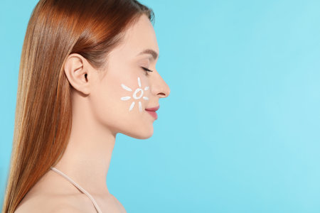 Beautiful young woman with sun protection cream on her face against light blue background, space for...