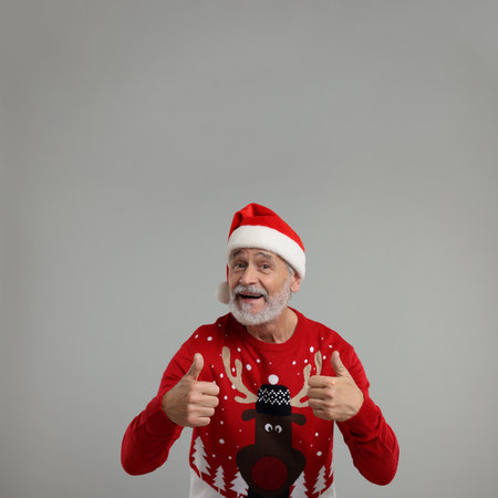 Senior man in Christmas sweater and Santa hat showing thumbs up on gray backgroundの写真素材