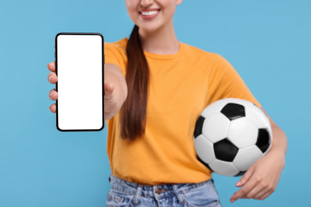 Happy soccer fan with ball showing smartphone on light blue background, closeupの写真素材