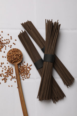 Uncooked buckwheat noodles (soba), grains and spoon on white tiled table, flat lay