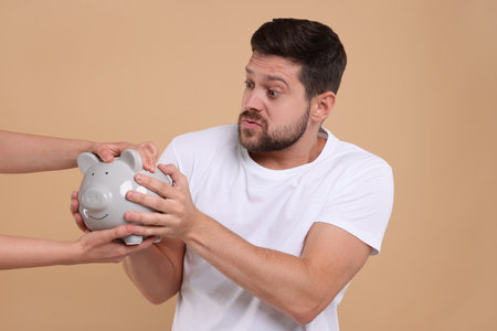 Woman taking piggy bank from emotional man on beige background. Be careful - fraudの写真素材