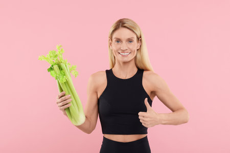 Happy woman with fresh green celery bunch showing thumb up against pink backgroundの写真素材