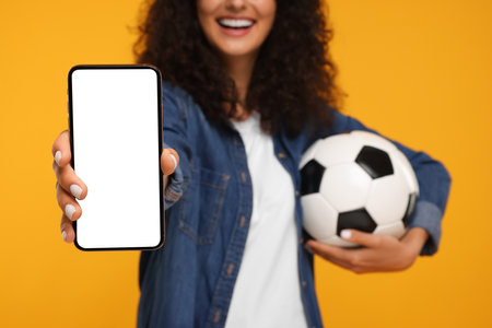 Happy fan holding soccer ball and showing smartphone on yellow background, closeupの写真素材
