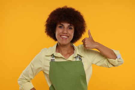 Happy young woman in apron showing thumb up on orange backgroundの写真素材