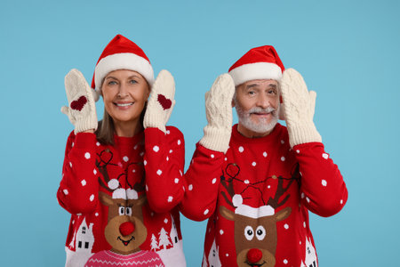 Happy senior couple in Christmas sweaters, Santa hats and knitted mittens on light blue backgroundの写真素材