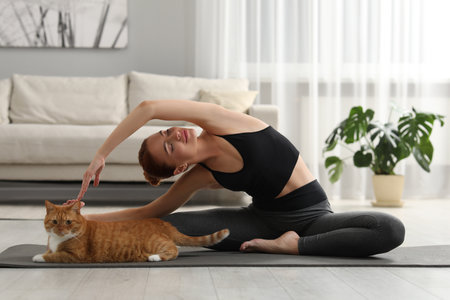 Beautiful woman with cute red cat practicing yoga on mat at homeの写真素材