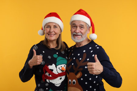 Happy senior couple in Christmas sweaters and Santa hats showing thumbs up on orange backgroundの写真素材
