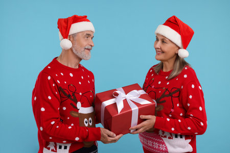 Senior couple in Christmas sweaters and Santa hats holding gift on light blue backgroundの写真素材