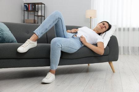 Young woman struggling to squeeze into tight jeans while lying on sofa at homeの写真素材