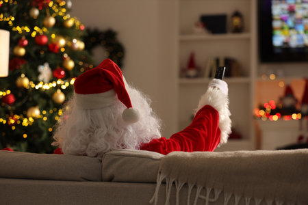 Merry Christmas. Santa Claus changing TV channels with remote control on sofa at home, back viewの写真素材