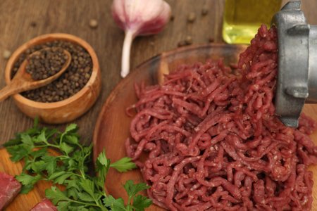 Meat grinder with beef mince, garlic, parsley and peppercorns on wooden table, closeup