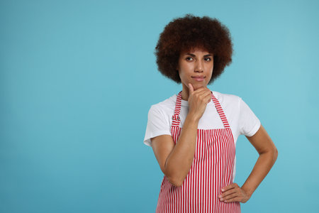 Thoughtful young woman in apron on light blue background. Space for textの写真素材