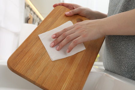 Woman wiping wooden cutting board with paper napkin at home, closeup