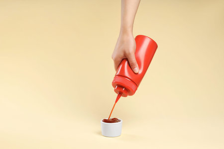 Woman squeezing tasty ketchup from bottle into bowl on beige background, closeupの写真素材